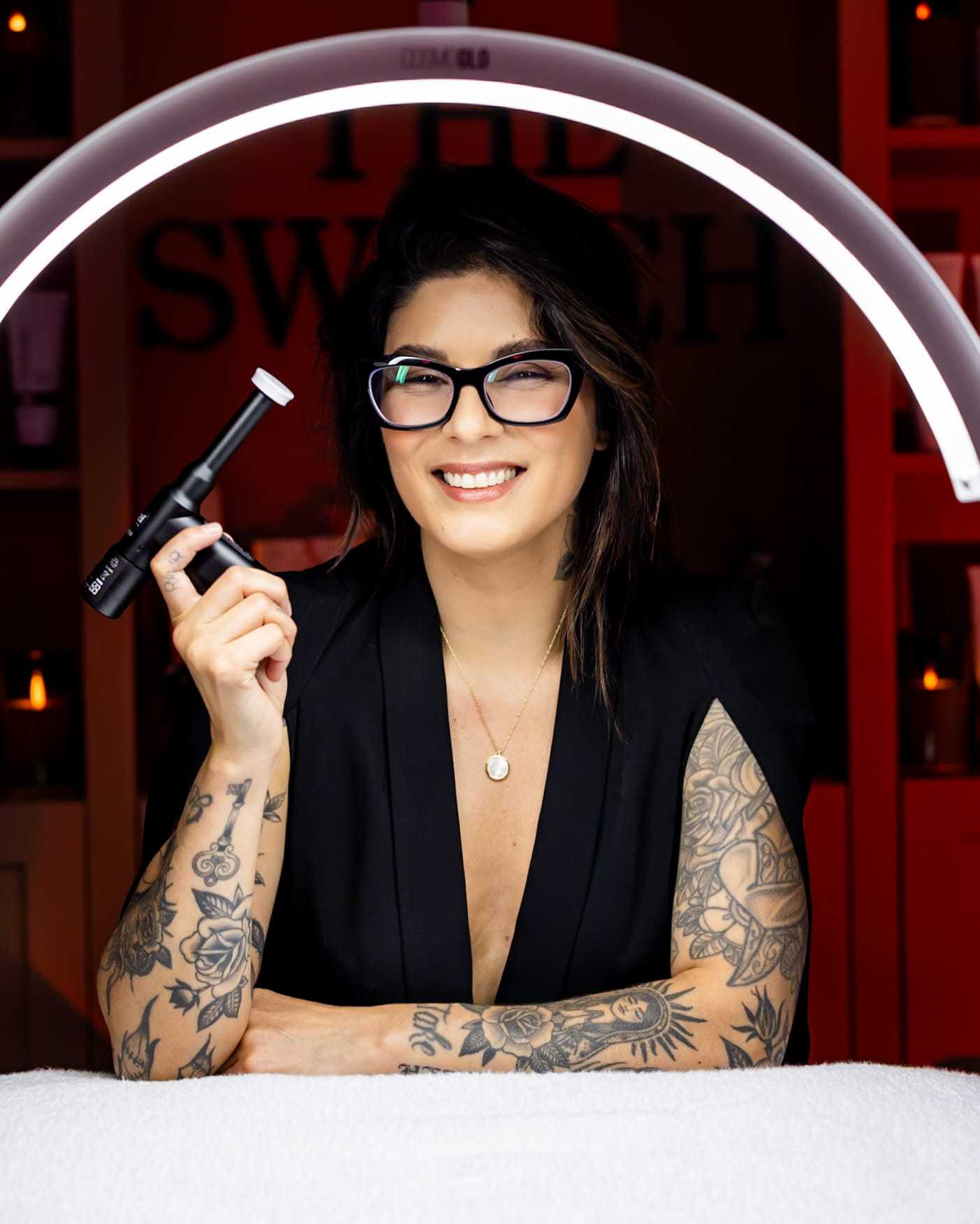 Woman with tattoos smiling, holding a black device under a ring light in a salon setting.