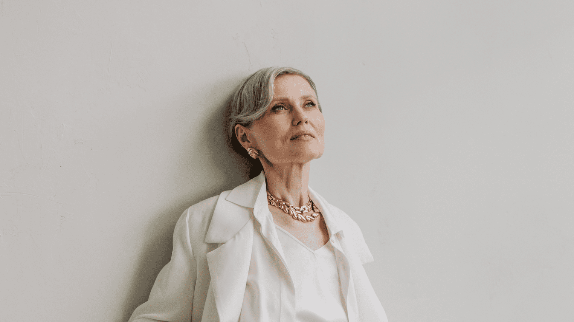 Mature woman in white outfit standing against a plain wall, gazing upwards.