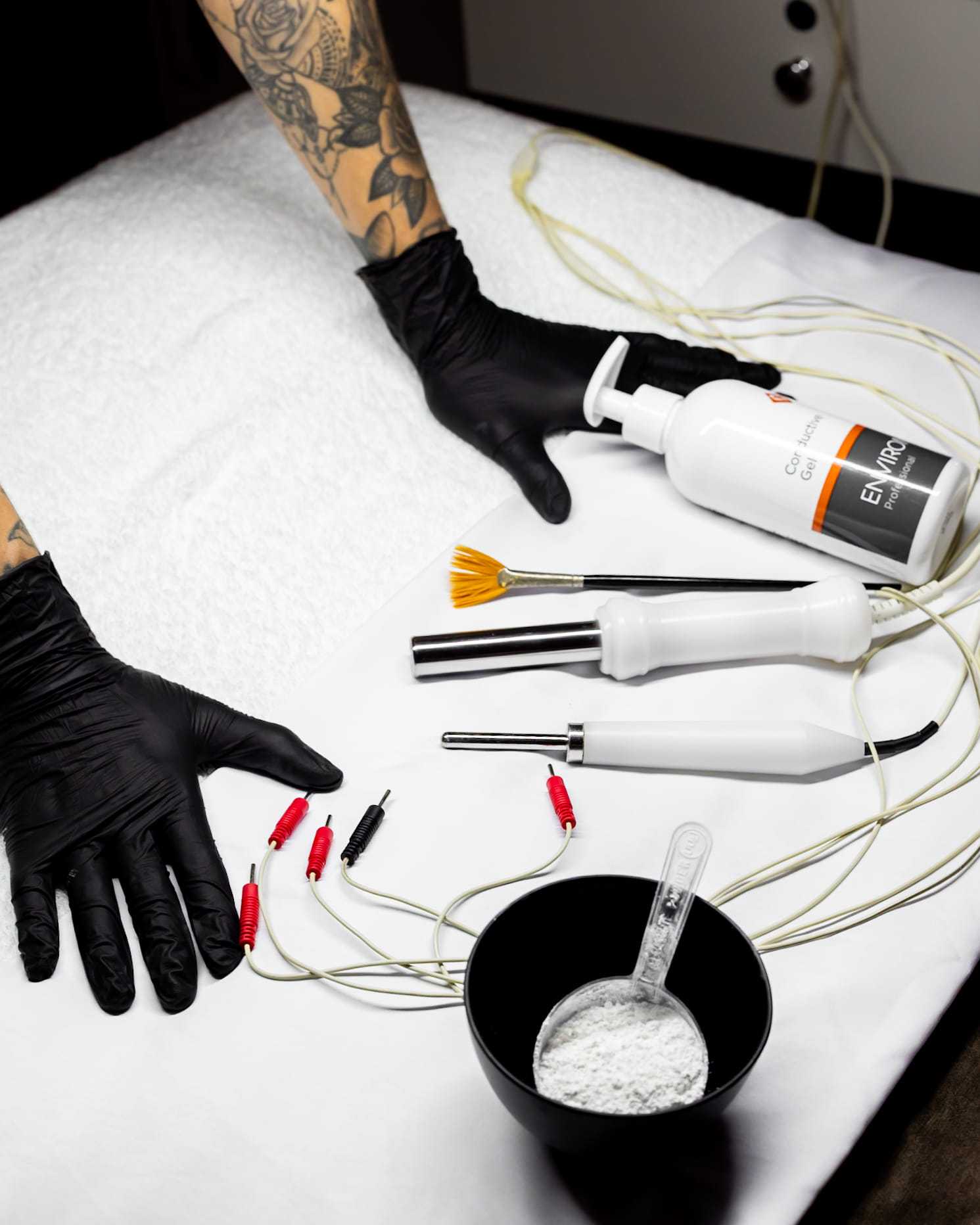 Tattooed arms with black gloves near skincare tools on a white surface.