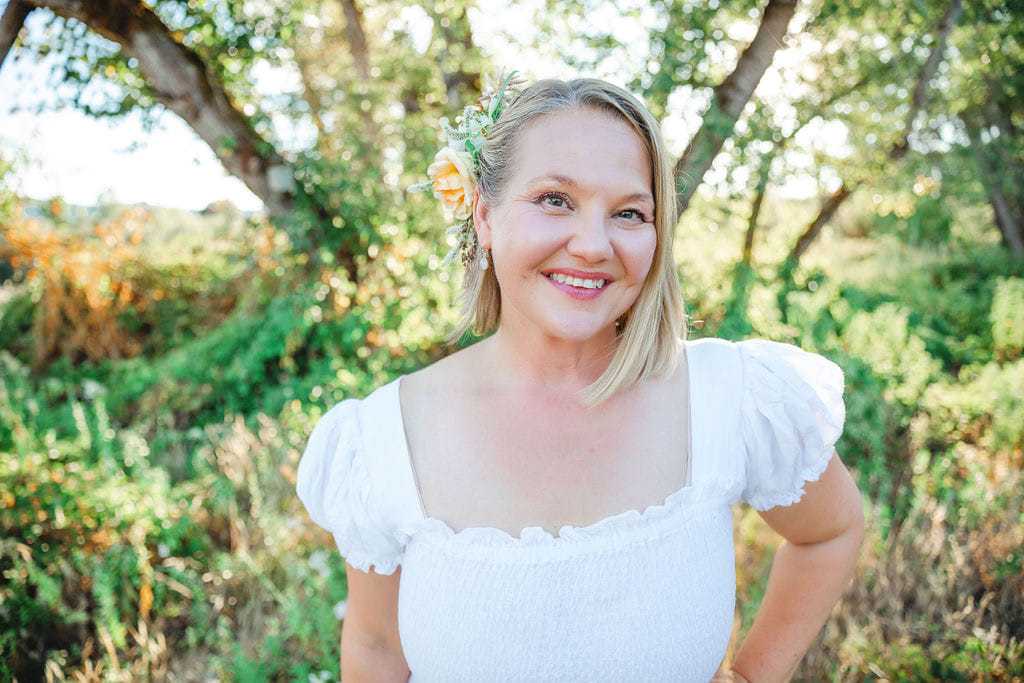 Woman standing outdoors, smiling, wearing a white dress with floral hair accessory.