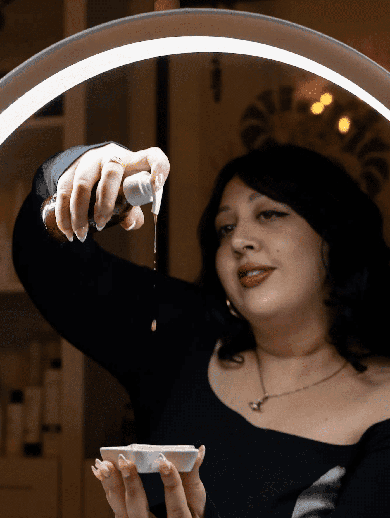 A person dispensing oil into a bowl under a ring light in a spa-like setting.